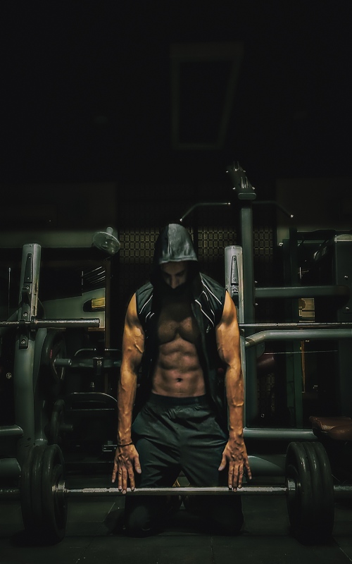 male bodybuilder wearing black sleeveless hoodie, opened from the front kneeling in a gym and gripping a barbell with weight plates.